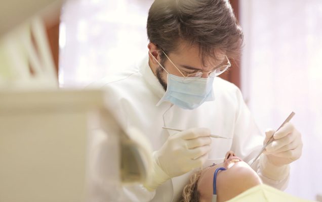 Dentysta w gabinecie zagląda do jamy ustnej pacjenta, w ręku trzyma narzędzia dentystyczne