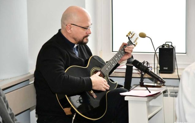 Msza święta w Wojskowym Instytucie Medycznym. Mężczyzna siedzi w ławce i gra na gitarze podczas mszy.