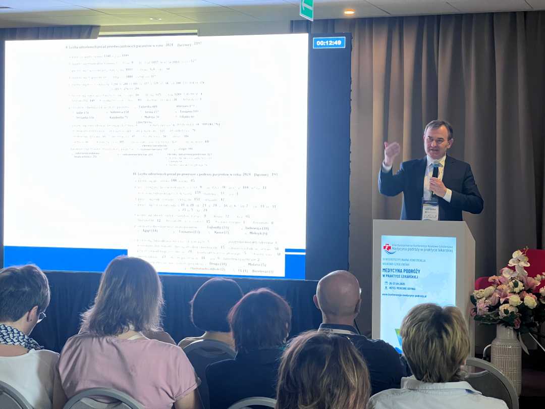 Wykładowca przy mównicy podczas konferencji. Po jego prawej stronie wyświetlony slajd z danymi statystycznymi.