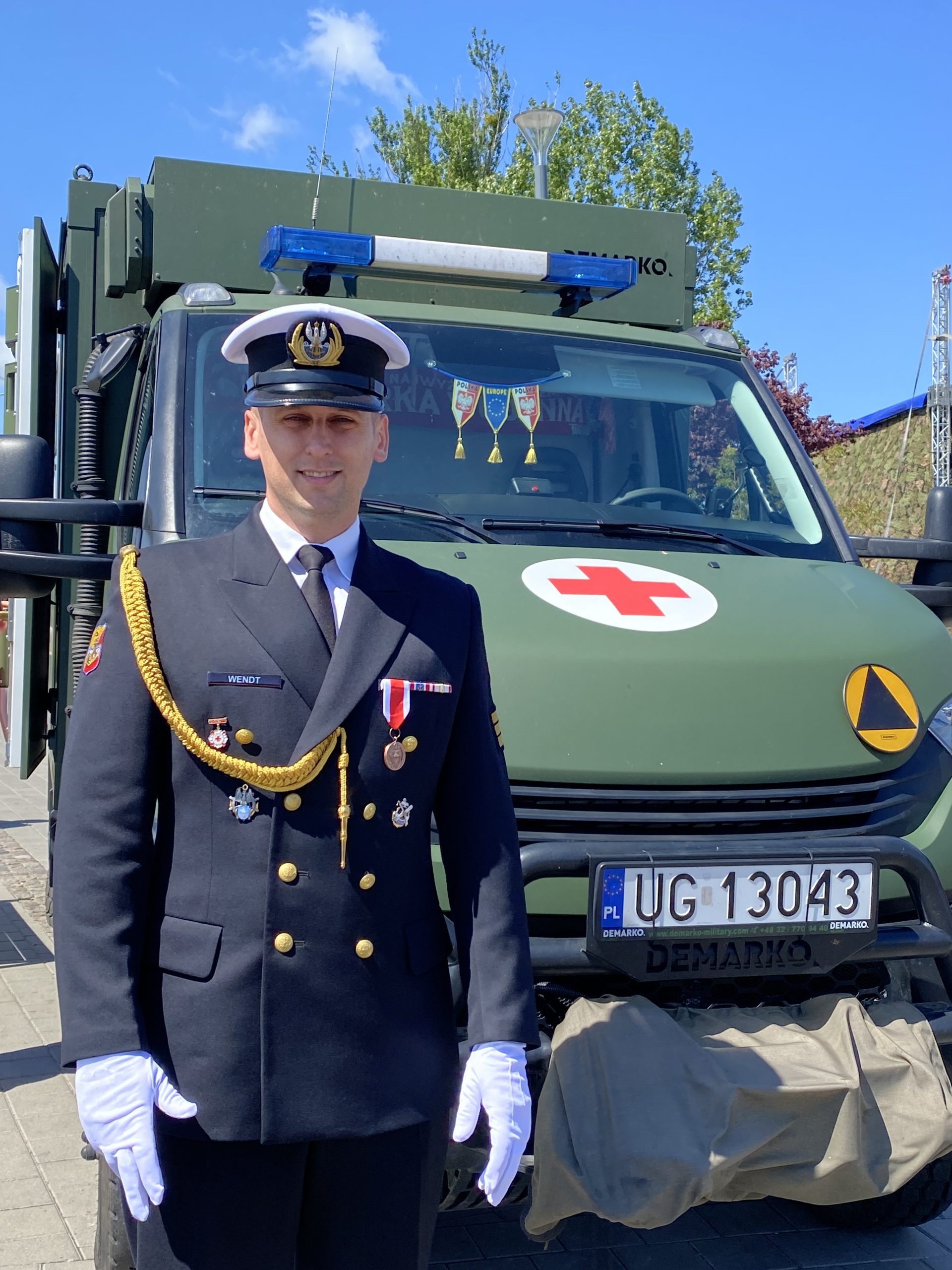 Żołnierz ubrany na galowo, za nim ambulans wojskowy.