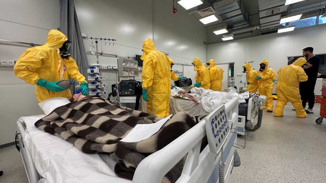 Medycy w żółtych kombinezonach i maskach przeciwgazowych podczas kursy CBRN w żółtych kombinezonach na sali szpitalnej.