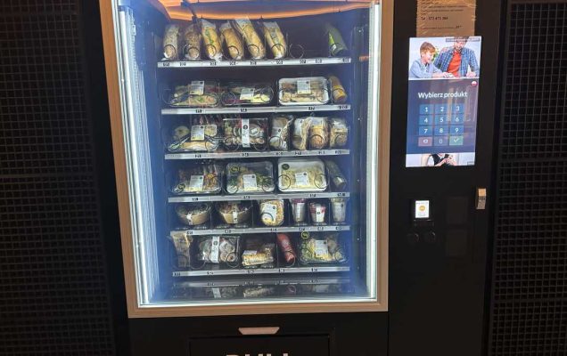 Automat do sprzedaży gotowych posiłków. Maszyna posiada duże szklane drzwi. Przez szybę widać na półkach zapakowane posiłki. Prawa część automatu zawiera wyświetlacz do składania zamówień oraz moduł do płatności.