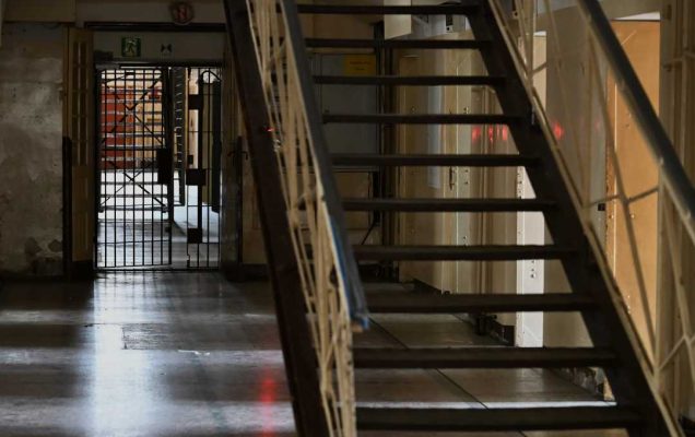 Korytarz w muzeum. Na pierwszym planie drewniane schody z prześwitem prowadzące w górę. Na drugim planie korytarz zakończony kratowymi stalowymi drzwiami.