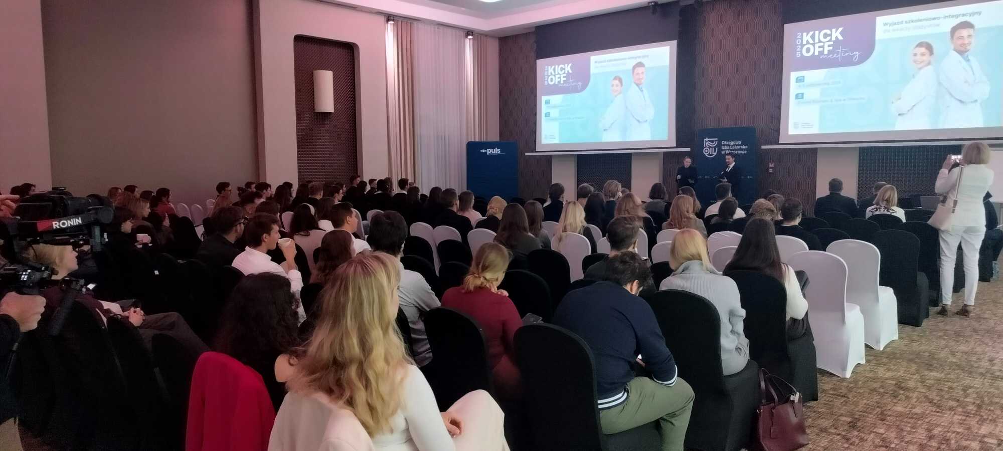 Ujęcie z tyłu sali. Uczestnicy konferencji na sali Konferencyjnej, na przeciw dwa ekrany wyświetlające materia konferencyjny.