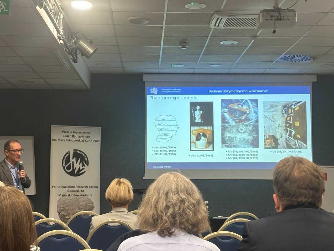 Sala wykładowa, ujęcie z tyłu sali uczestników siedzących na sali konferencyjnej podczas wykładu. Przed siedzącymi stojący prelegent przy prezentacji wyświetlanej z rzutnika.