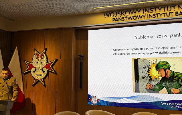 Panelista przy mównicy, za nim logo WIM i flagi Polski, od lewej strony na ekranie fragment prezentacji na ekranie.