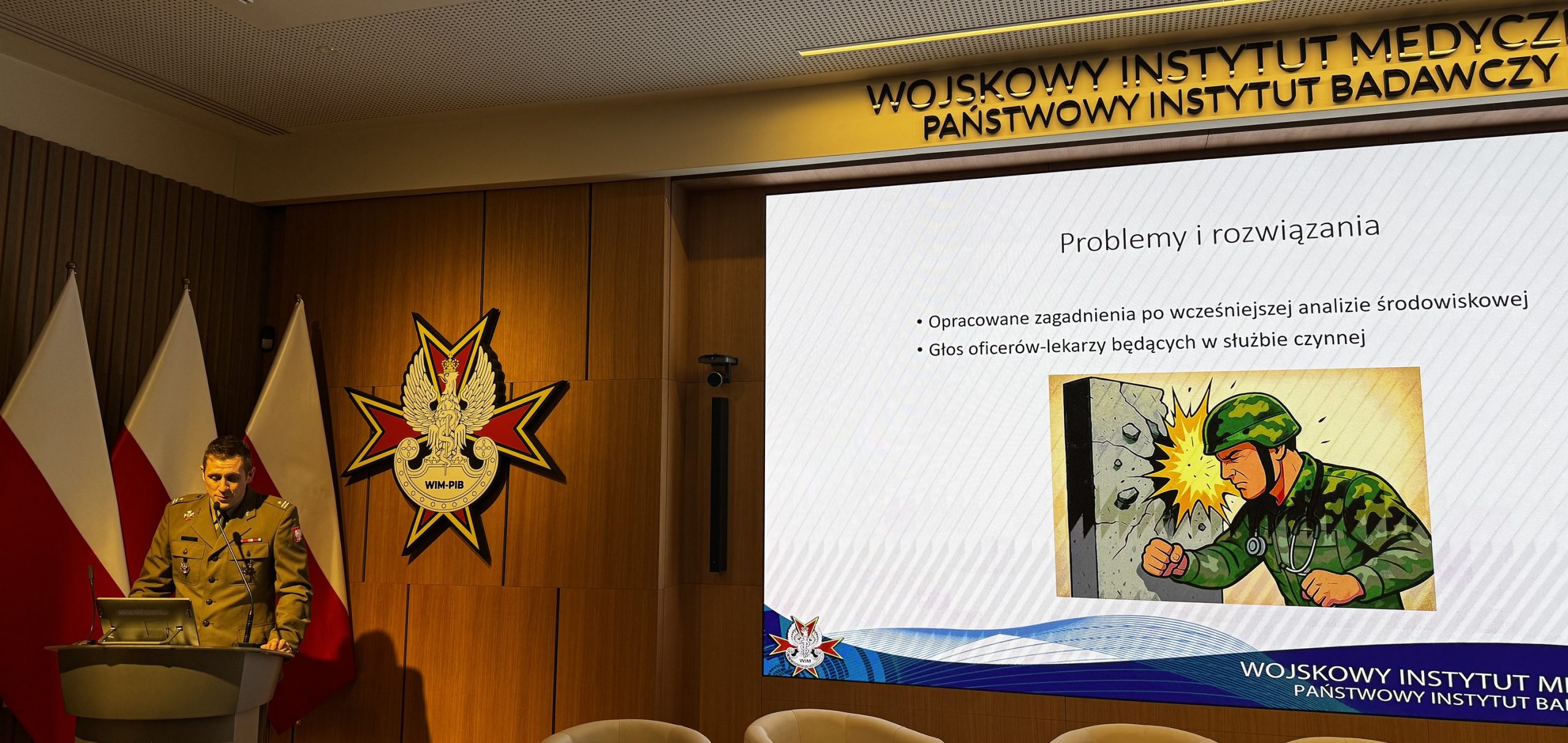 Panelista przy mównicy, za nim logo WIM i flagi Polski, od lewej strony na ekranie fragment prezentacji na ekranie.