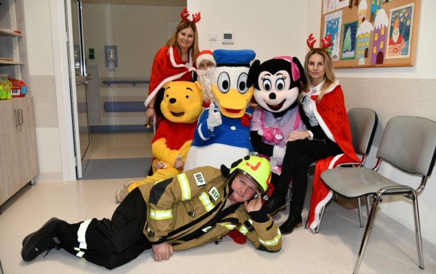 Na szpitalnym korytarzu pozują pomocnicy świętego mikołaja. Na podłodze leży Strażak sam, obok niego siedzą Kubuś Puchatek, Myszka Miki, Kaczor Donald oraz dwie śnieżynki.