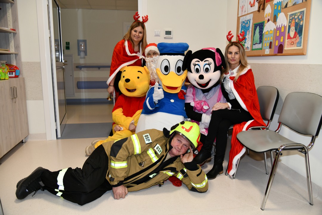 Na szpitalnym korytarzu pozują pomocnicy świętego mikołaja. Na podłodze leży Strażak sam, obok niego siedzą Kubuś Puchatek, Myszka Miki, Kaczor Donald oraz dwie śnieżynki.