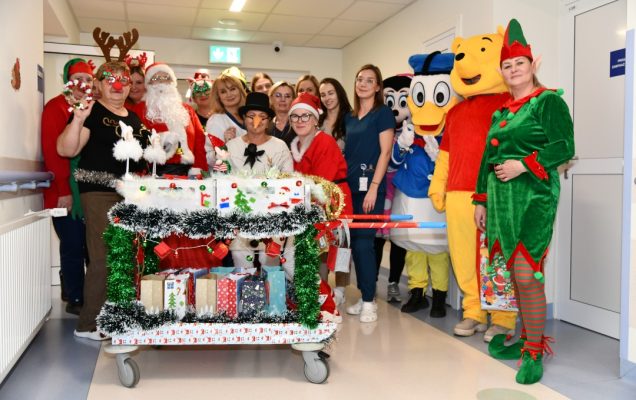 Pomocnicy świętego Mikołaja na korytarzu szpitalnym z ozdobionym wózkiem pełnym prezentów.