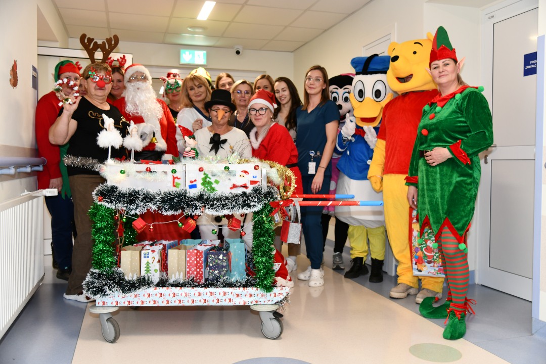 Pomocnicy świętego Mikołaja na korytarzu szpitalnym z ozdobionym wózkiem pełnym prezentów.