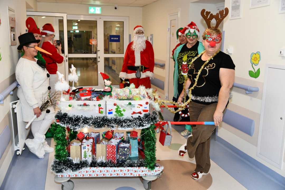 Pomocnicy świętego Mikołaja na korytarzu szpitalnym z ozdobionym wózkiem pełnym prezentów.