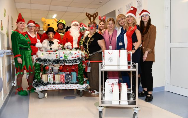 Pomocnicy świętego Mikołaja na korytarzu szpitalnym z ozdobionym wózkiem pełnym prezentów.