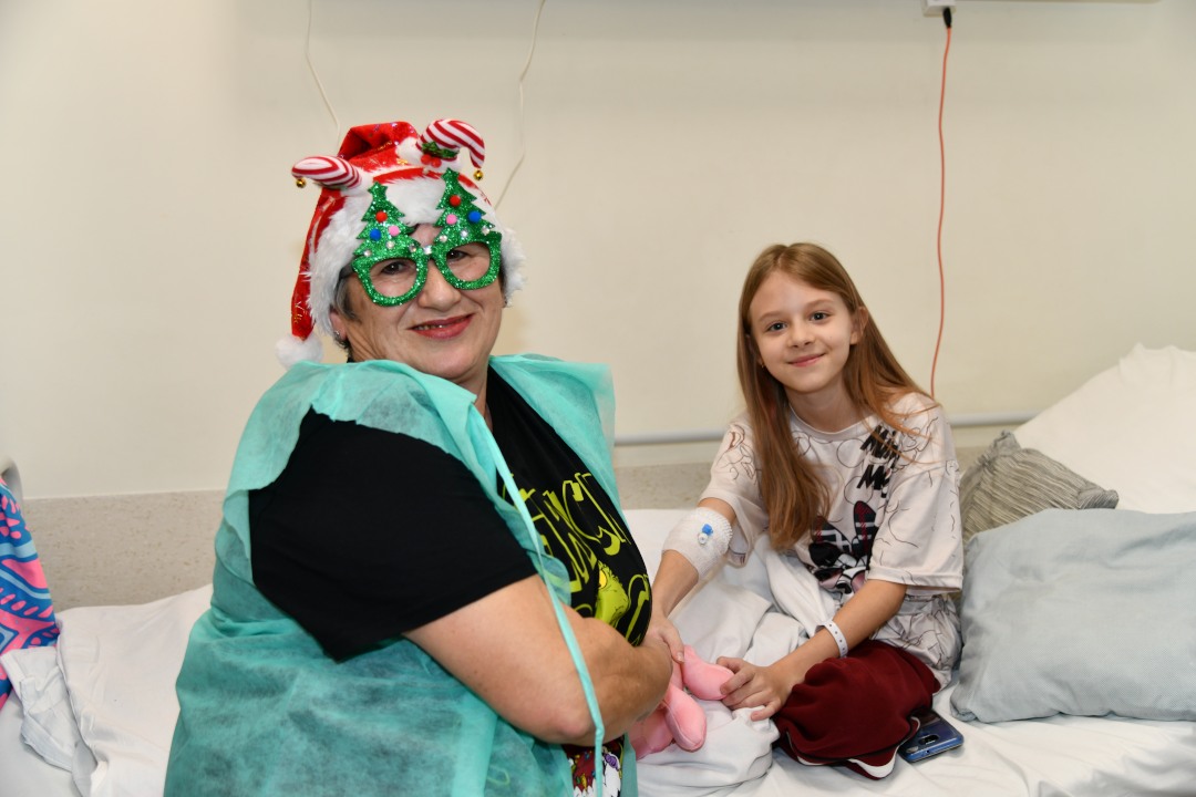 Pomocniczka świętego Mikołaja z kilkuletnią pacjentką siedzą na łóżku szpitalnym.