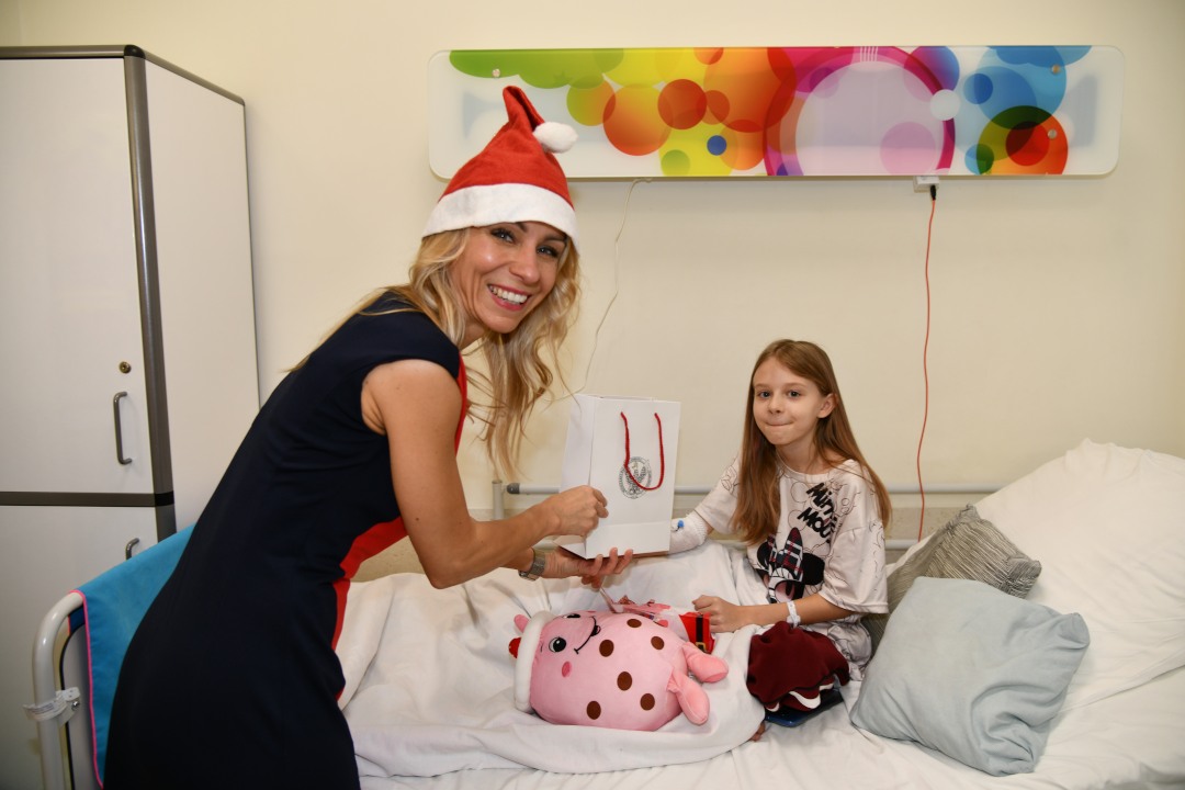 Sala szpitalna. Małgorzata Złotkowska z czapką świętego Mikołaja na głowie wręcza prezent małej pacjentce.