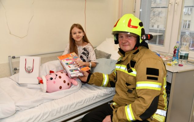Sala szpitalna. Pan strażak wręcza prezent małej pacjentce.