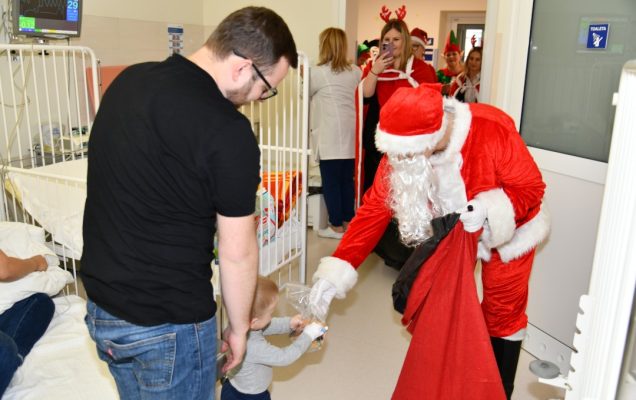 Sala szpitalna, święty Mikołaj pochyla się i wręcza 2 letniemu pacjentowi prezent. Za chłopcem stoi jego tata.