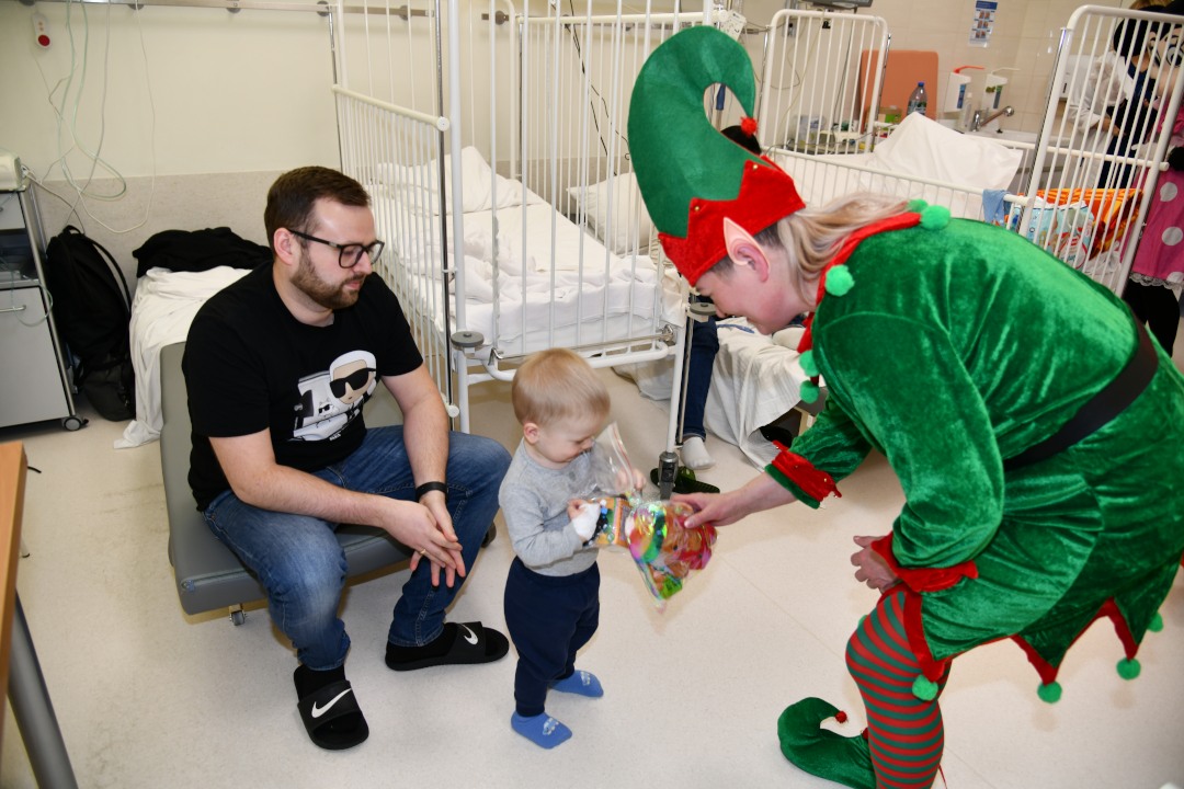 Sala szpitalna, Pani Elfowa pochyla się i wręcza 2 letniemu pacjentowi prezent. Za chłopcem kuca jego tata.