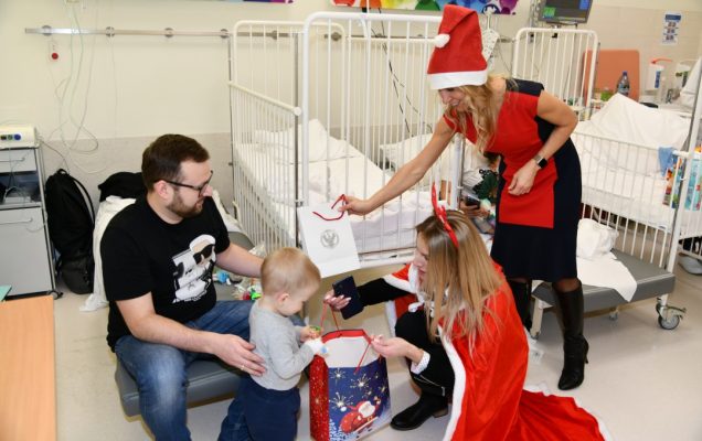 Sala szpitalna, Małgorzata Złotkowska pochyla się i wręcza 2 letniemu pacjentowi prezent. Za chłopcem stoi jego tata.