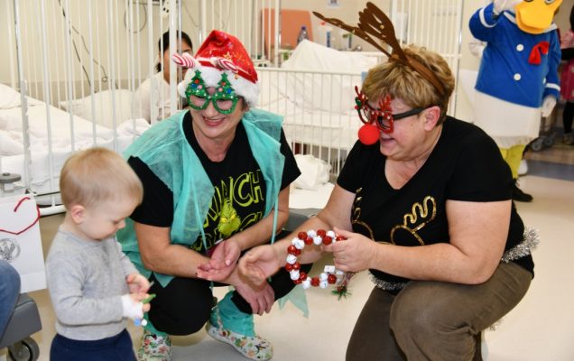 Pomocniczki świętego Mikołaja kucają i wręczają prezent maleńkiemu chłopcu.