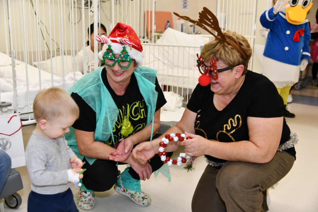 Pomocniczki świętego Mikołaja kucają i wręczają prezent maleńkiemu chłopcu.