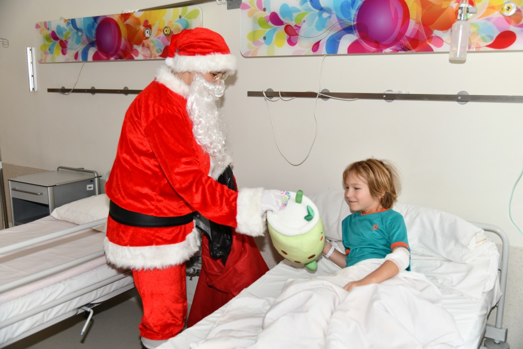 Sala szpitalna. Święty Mikołaj przy łóżku wręcza prezent młodemu pacjentowi.