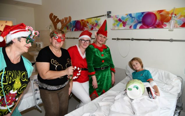 Sala szpitalna. Pomocnicy Świętego Mikołaja pozują z młodym pacjentem leżącym na łóżku do zdjęcia.