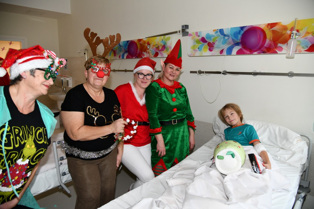 Sala szpitalna. Pomocnicy Świętego Mikołaja pozują z młodym pacjentem leżącym na łóżku do zdjęcia.