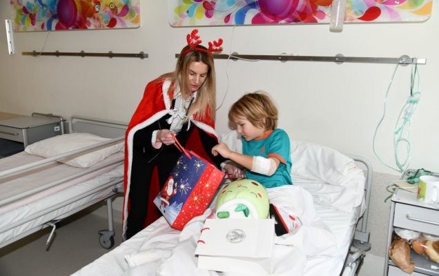 Sala szpitalna. Mały pacjent w łóżku szpitalnym przy pomocy śnieżynki otwiera torbę z prezentami.