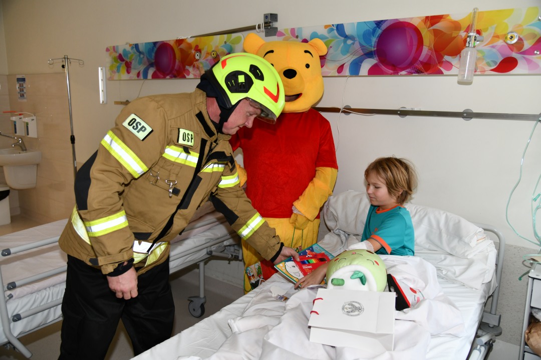 Sala szpitalna. Mały pacjent w łóżku szpitalnym ze strażakiem i Kubusiem Puchatkiem oglądają prezenty.