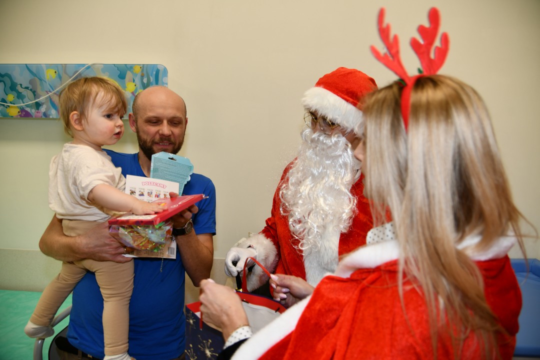 Święty Mikołaj i jego pomocnicy odwiedzają salę najmłodszych pacjentów. Tata z małym dzieckiem na ręku.