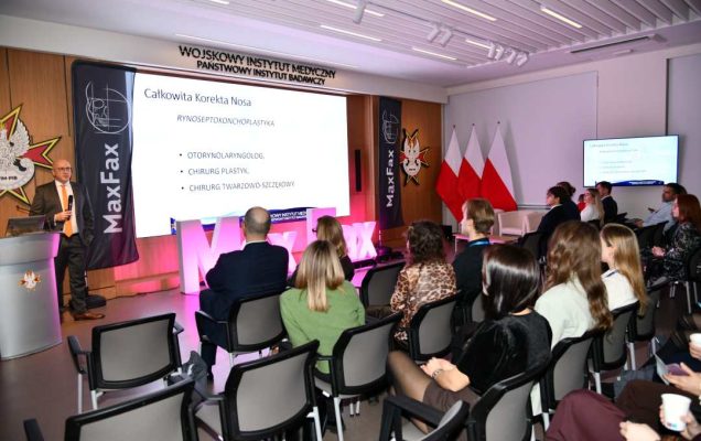 Siedzący słuchacze słuchający stojącego przed nimi prelegenta. Za prelegentem slajd z konferencji.