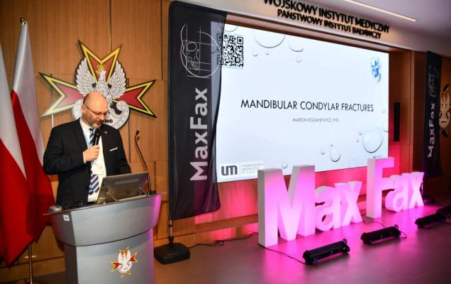 Prelegent przy mównicy. Obok niego stojący różowy napis Max Fax i ekran ze slajdem z konferencji.