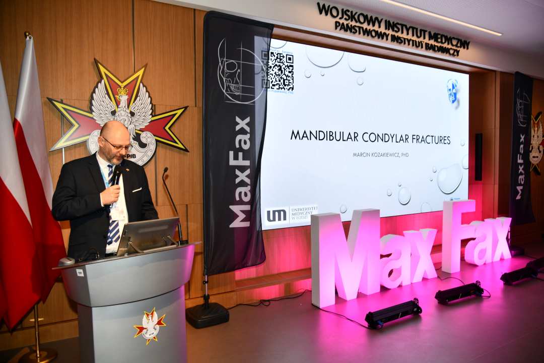 Prelegent przy mównicy. Obok niego stojący różowy napis Max Fax i ekran ze slajdem z konferencji.