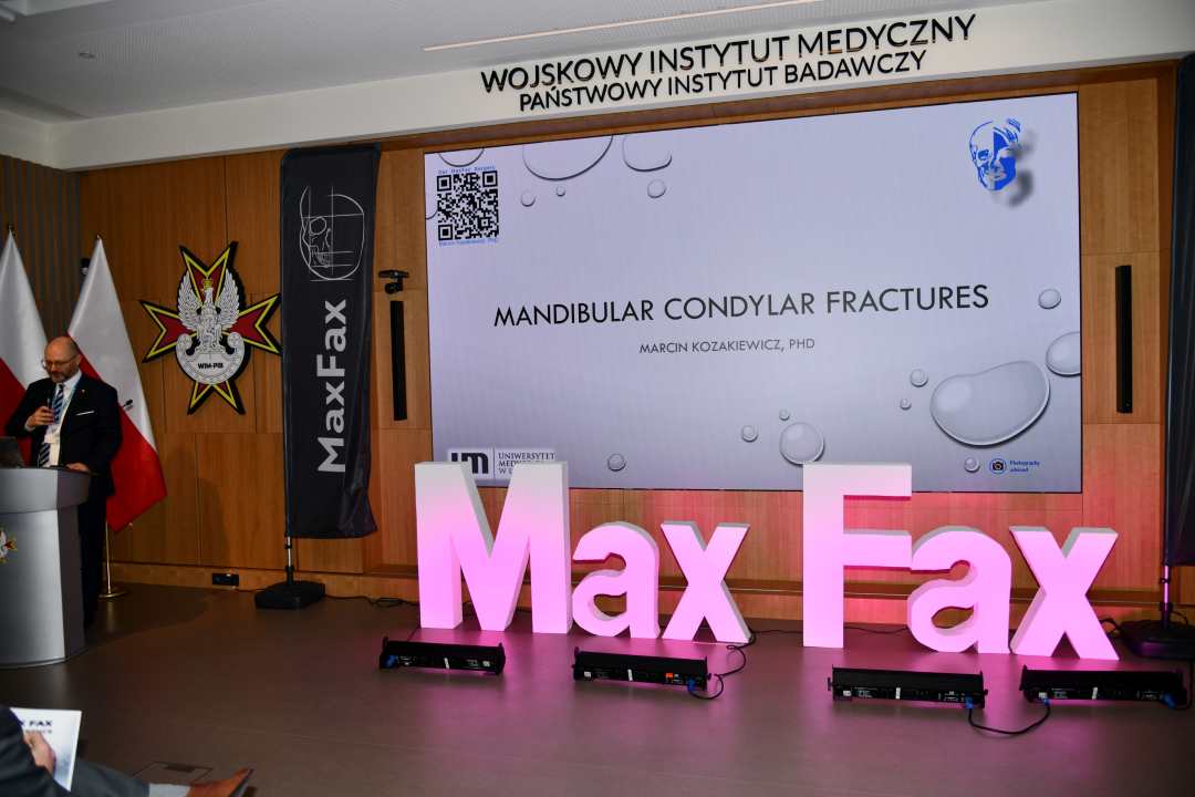 Prelegent przy mównicy. Obok niego stojący różowy napis Max Fax i ekran ze slajdem z konferencji.