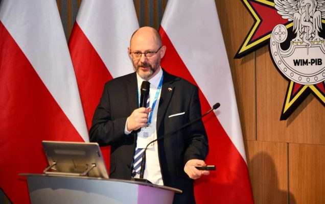 Prelegent przy mównicy, za nim logo WIM i stojące 3 flagi Polski.