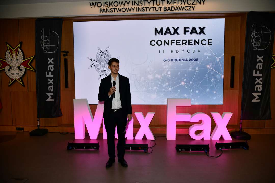 Prelegent podczas konferencji. Za nim podświetlony napis Max Fax i slajd tytułowy konferencji MAxFax.