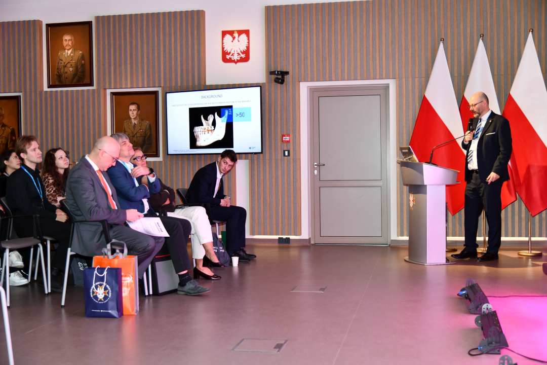 Prelegent przy mównicy mówiący do słuchaczy. Za nim logo WIM i stojące 3 flagi Polski.
