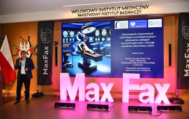 Prelegent przy mównicy. Za nim logo WIM. Obok stoi różowa dekoracja z napisem Max Fax i ekran z wyświetlonym slajdem z konferencji.