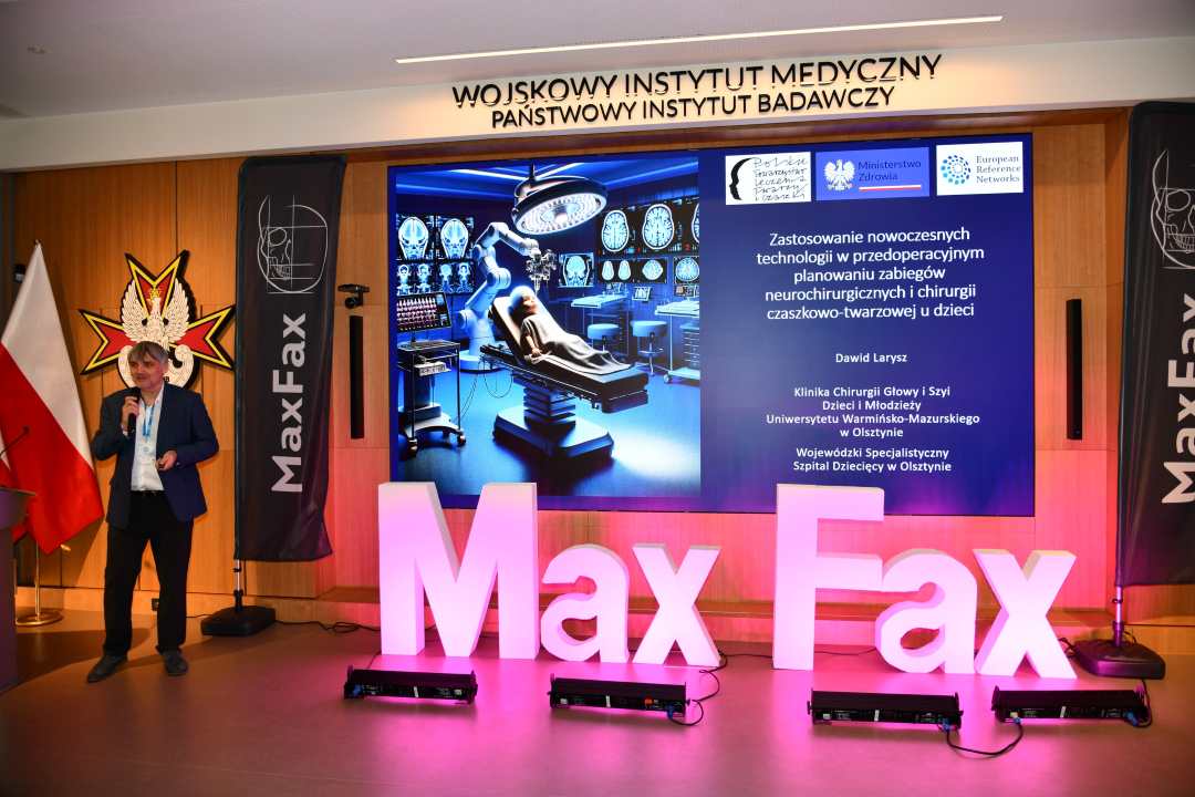 Prelegent przy mównicy. Za nim logo WIM. Obok stoi różowa dekoracja z napisem Max Fax i ekran z wyświetlonym slajdem z konferencji.