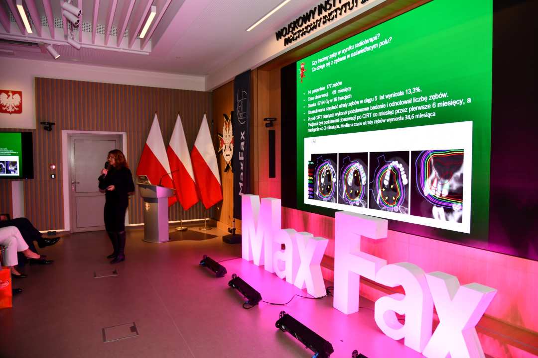 Prelegentka prowadząca wykład. Za nią logo WIM. Obok stoi różowa dekoracja z napisem Max Fax i ekran z wyświetlonym slajdem z konferencji.