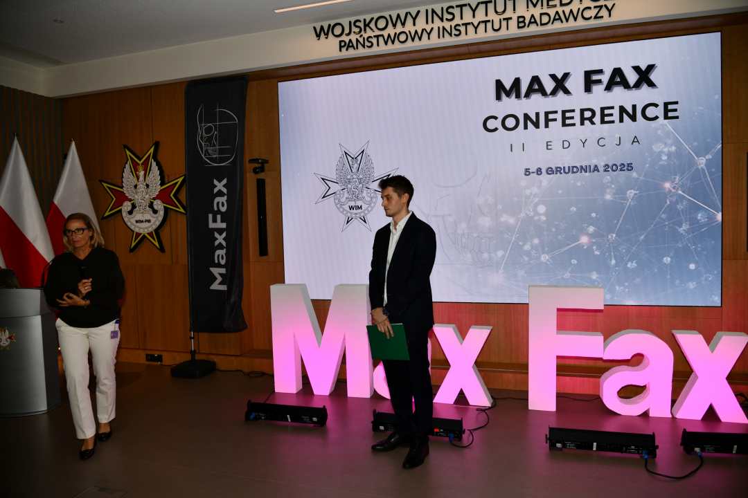 Prelegent podczas konferencji. Za nim podświetlony napis Max Fax i slajd tytułowy konferencji MAxFax. Po prawej stronie doktor Aldona Chloupek.