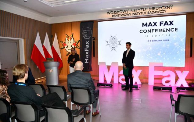 Prelegent podczas konferencji. Za nim podświetlony napis Max Fax i slajd tytułowy konferencji MAxFax.