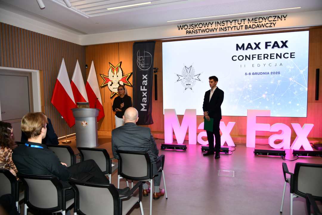 Prelegent podczas konferencji. Za nim podświetlony napis Max Fax i slajd tytułowy konferencji MAxFax.