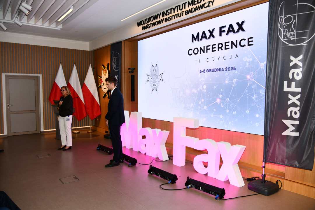 Prelegent podczas konferencji. Za nim podświetlony napis Max Fax i slajd tytułowy konferencji MAxFax.