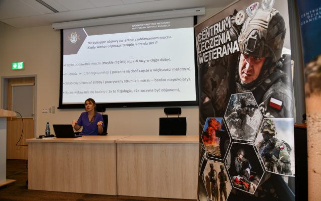 ppłk dr n. med. Agnieszka Grabińska prowadzi wykład. Za jej plecami plansza wyświetlanej prezentacji. Z boku Stojak z plakatem Centrum leczenie weterana, na plakacie kolaż zdjęć z akcji wojskowych.