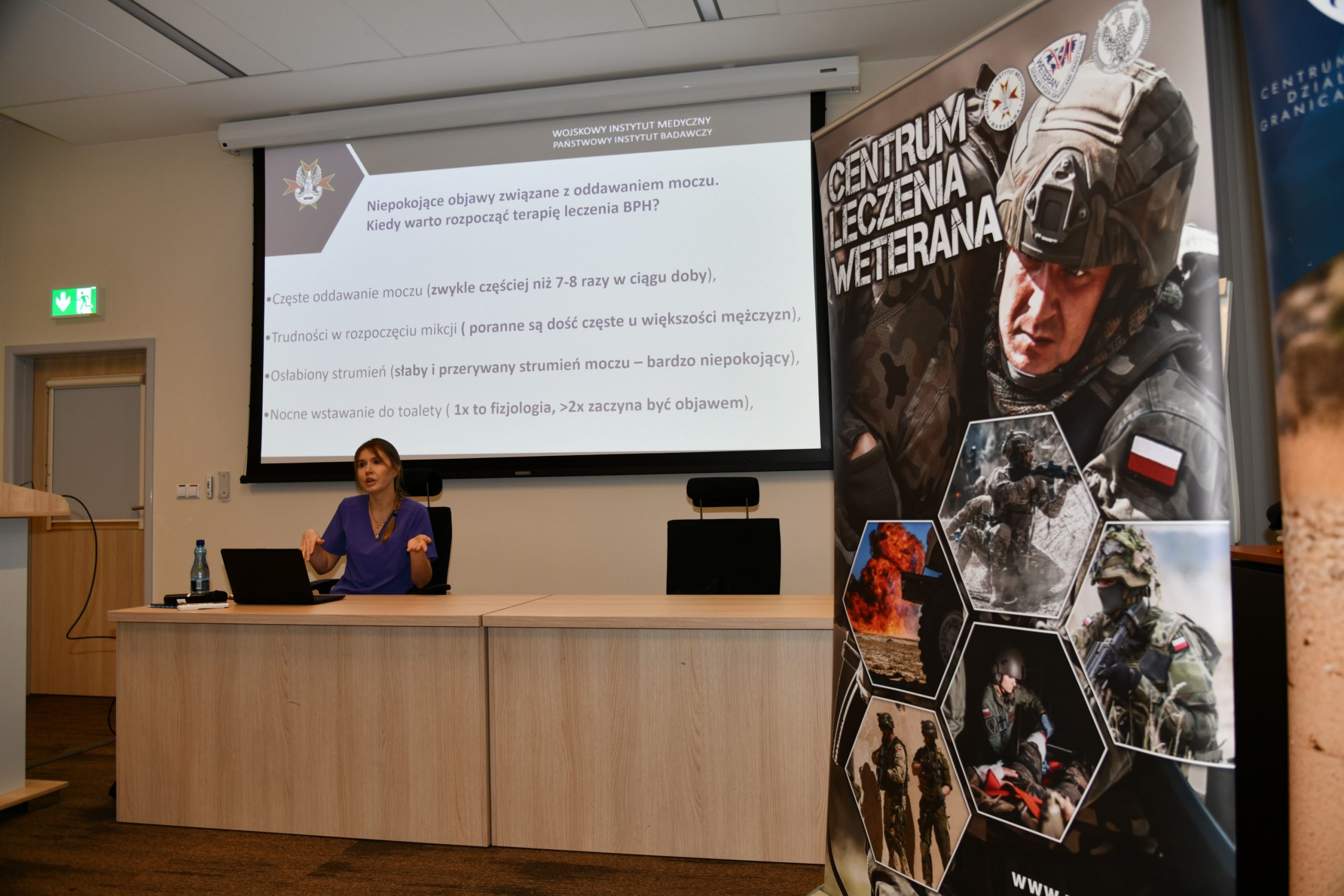 ppłk dr n. med. Agnieszka Grabińska prowadzi wykład. Za jej plecami plansza wyświetlanej prezentacji. Z boku Stojak z plakatem Centrum leczenie weterana, na plakacie kolaż zdjęć z akcji wojskowych.