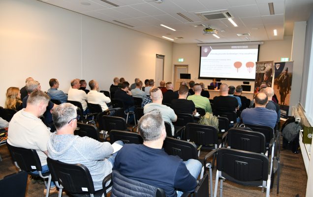 Ujęcie z tyłu na siedzących słuchaczy podczas wykładu. Ppłk dr n. med. Agnieszka Grabińska prowadzi wykład. Za jej plecami plansza wyświetlanej prezentacji. Z boku Stojak z plakatem Centrum leczenie weterana, na plakacie kolaż zdjęć z akcji wojskowych.