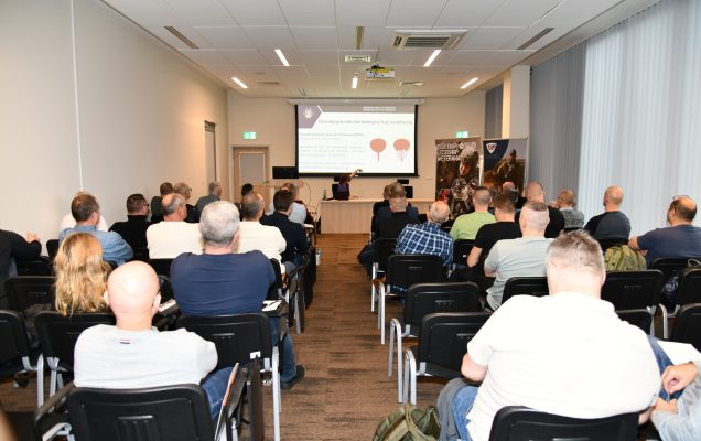 Ujęcie z tyłu na siedzących słuchaczy podczas wykładu. Ppłk dr n. med. Agnieszka Grabińska prowadzi wykład. Za jej plecami plansza wyświetlanej prezentacji. Z boku Stojak z plakatem Centrum leczenie weterana, na plakacie kolaż zdjęć z akcji wojskowych.