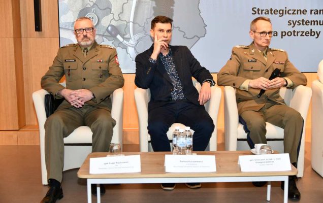 Generał broni Grzegorz Gielerak siedzący z panelistami podczas sesji, za nimi fragment wyświetlanej prezentacji.